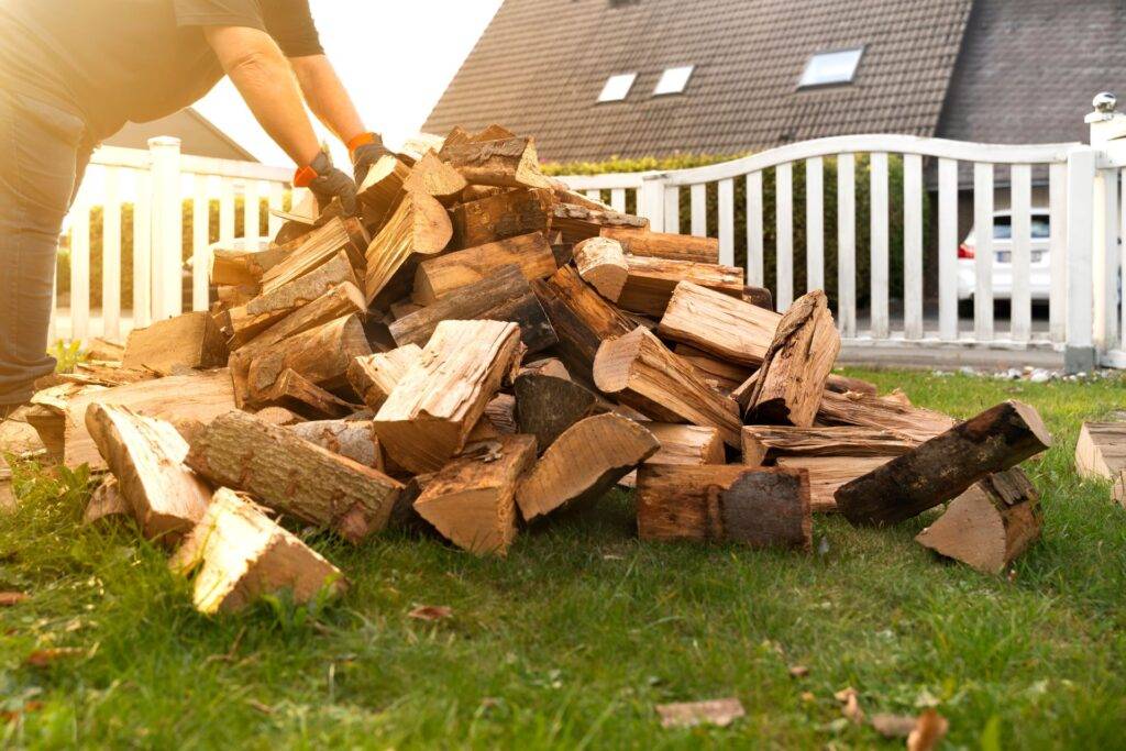 Im Garten liegt ein Stapel Buchenscheite, die angeliefert wurden. Ein Mann bückt sich und hebt einen Arm voll Scheite hoch um ihn ins Kaminholzlager zu bringen. Herbstabend mit Sonnenlicht